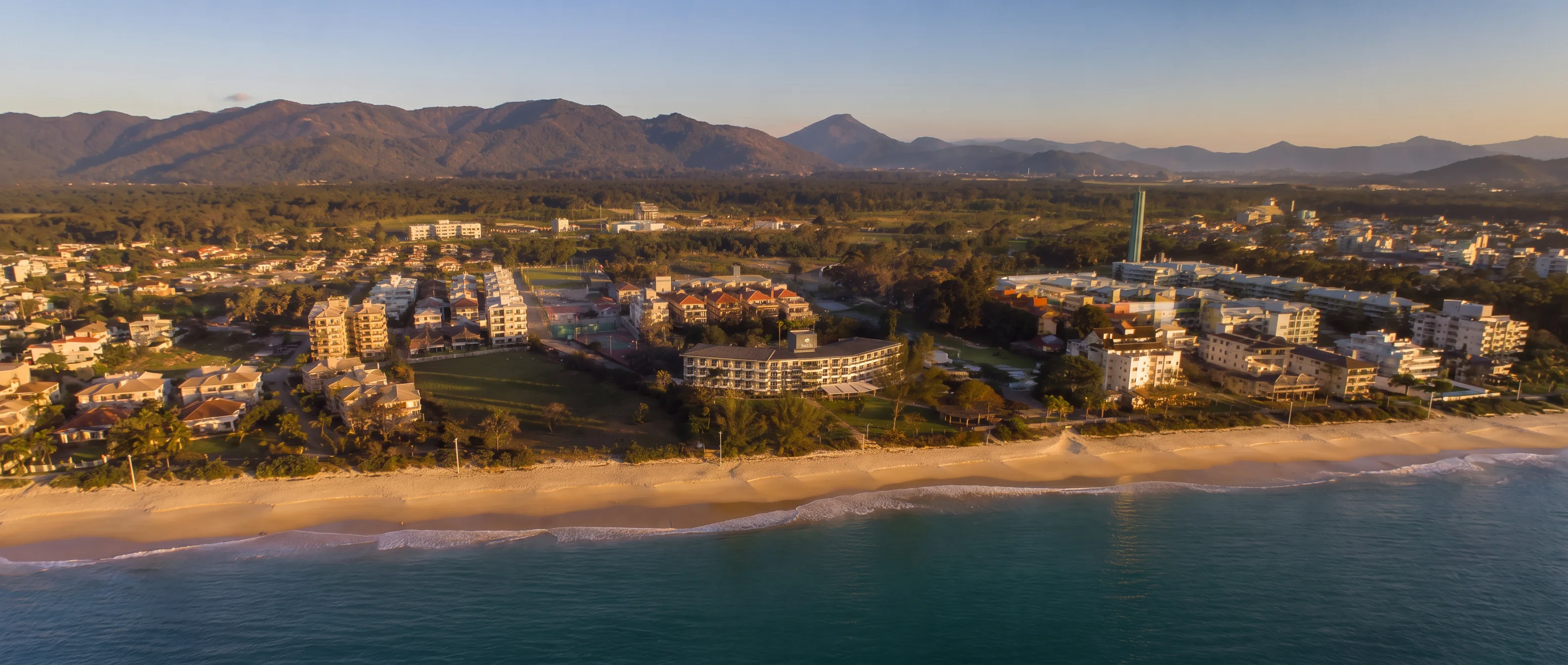 Vista aérea de Cachoeira do Bom Jesus ao pôr do sol: o Hotel Porto Sol Beach à beira-mar, a praia de Canasvieiras, o mar e as montanhas ao fundo